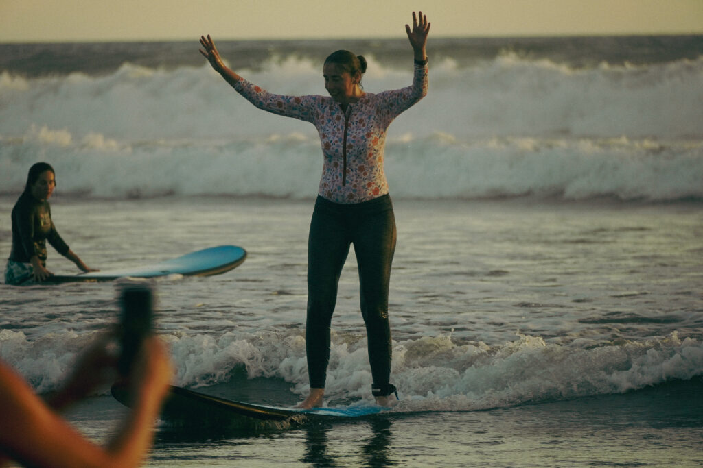 Woman standing on a surfboard with arms raised at the Pura Vida Adventures Women 50+ surf retreat in Santa Teresa, Costa Rica